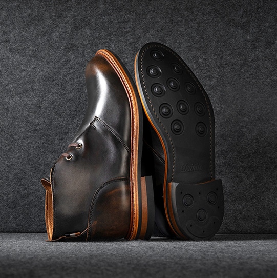 A pair of brown dress shoes with a burnished finish, shown on a dark background. One shoe is displayed upright and the other lying on its side to reveal the polished leather sole.