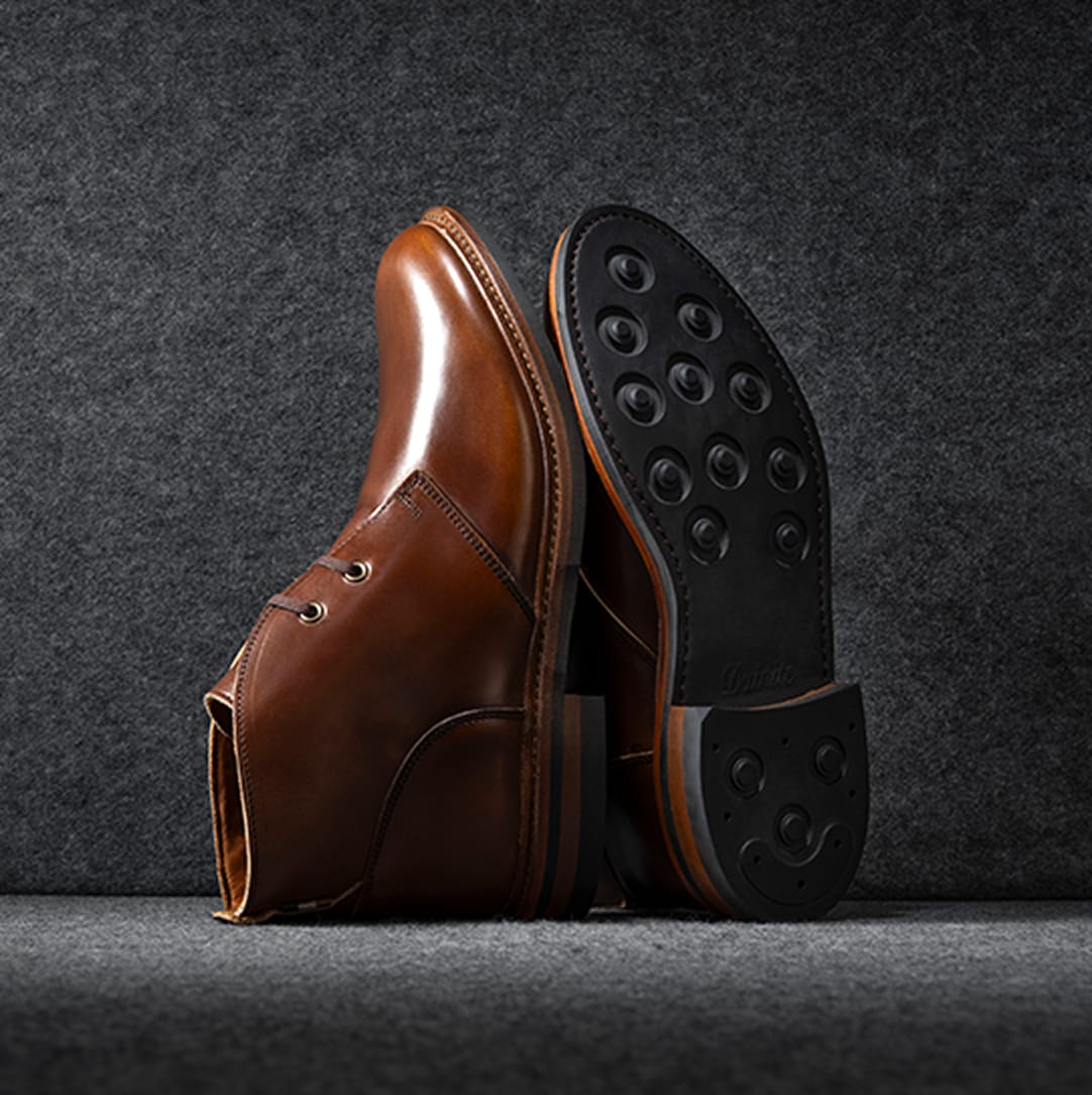 A pair of brown leather Chukka boots displayed vertically, showing the black dainite leather sole.