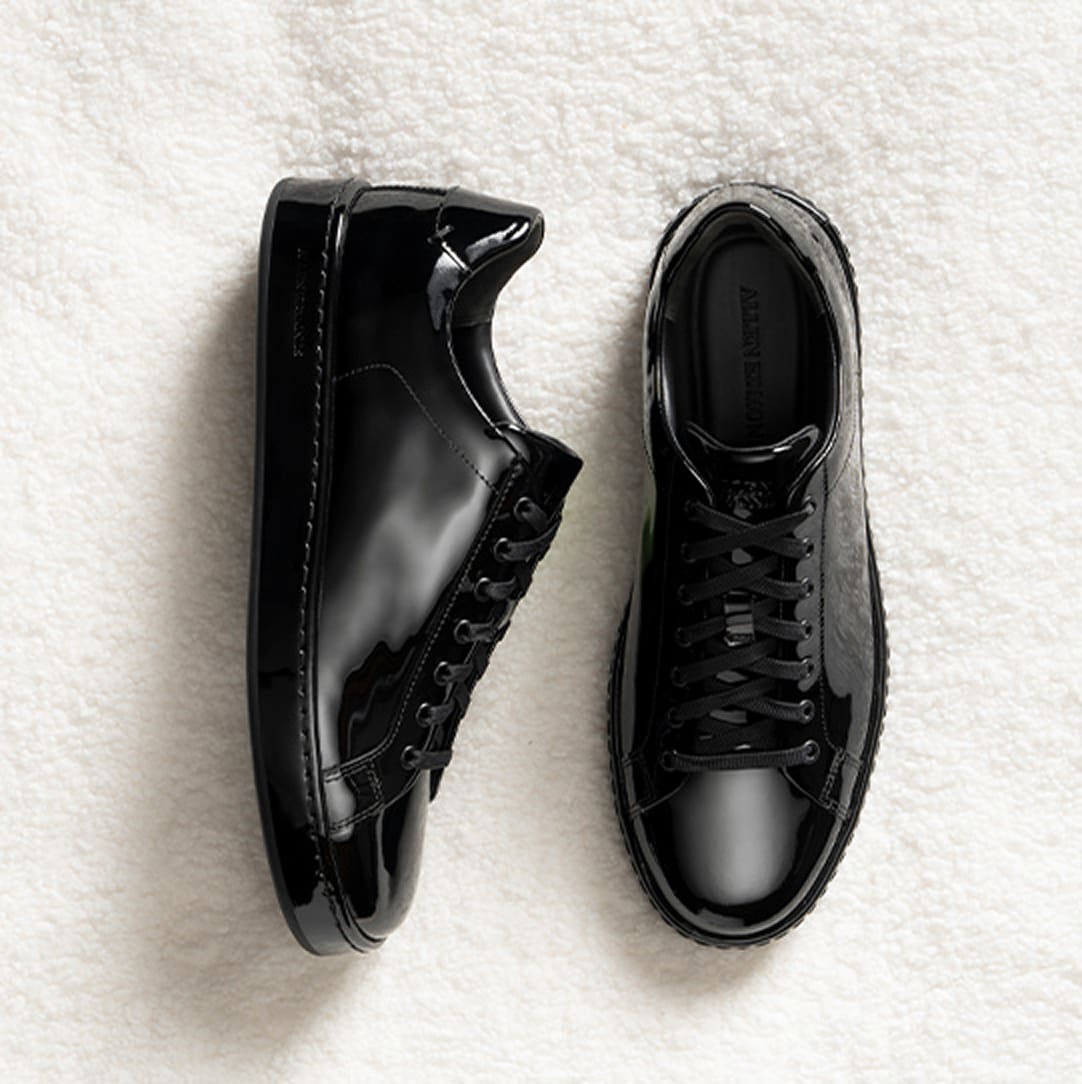 A pair of all-black patent leather sneakers with black laces, with one shoe lying on its side and the other facing upright.