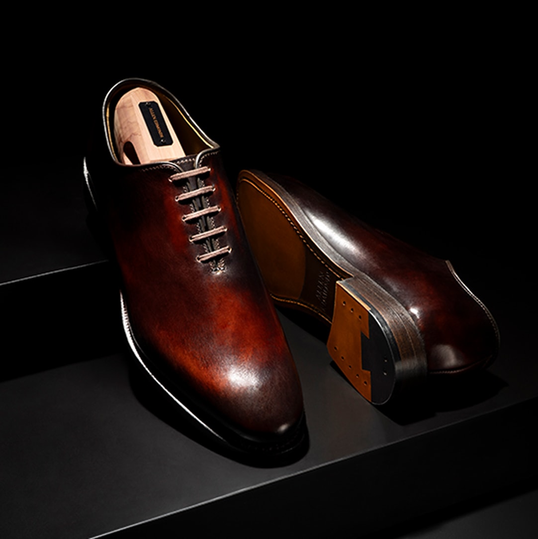 A pair of brown dress shoes with a burnished finish, shown on a dark background. One shoe is displayed upright and the other lying on its side to reveal the polished leather sole.