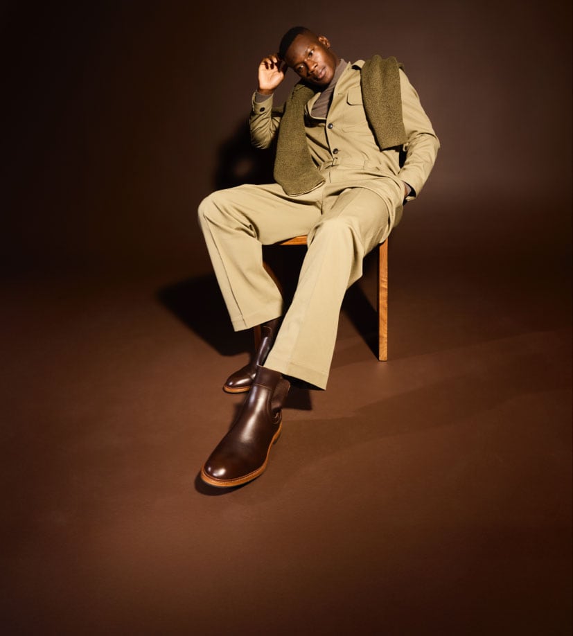 A man sitting in a chair modeling a tan pair of pants and matching jacket, with a green sweater draped over his shoulder and a pair of brown leather Chelsea boots.