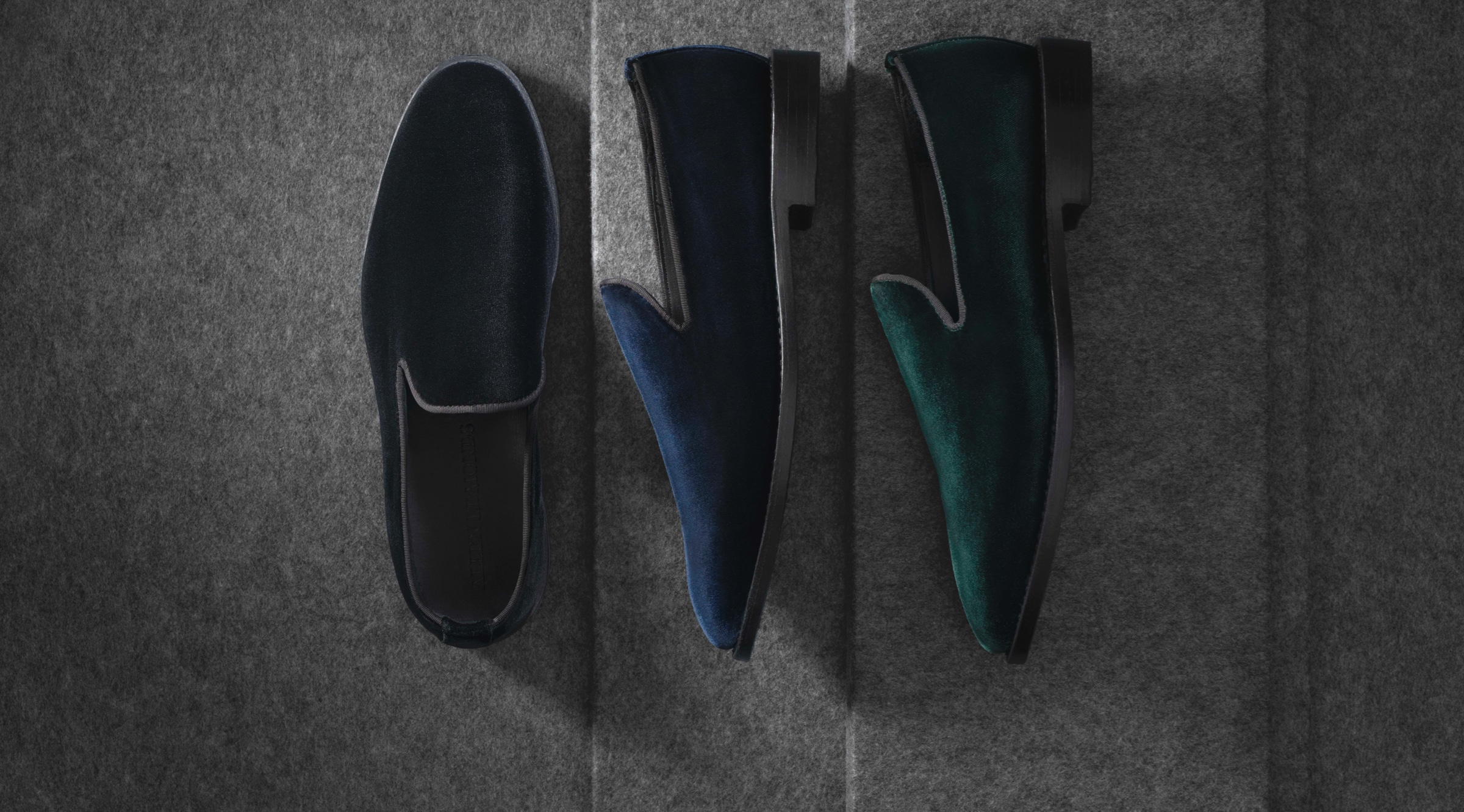 A top view of three velvet loafers. One is black, one is blue, and one is green. The black one is displayed on its sole, while the green and blue shoes are displayed on their side.