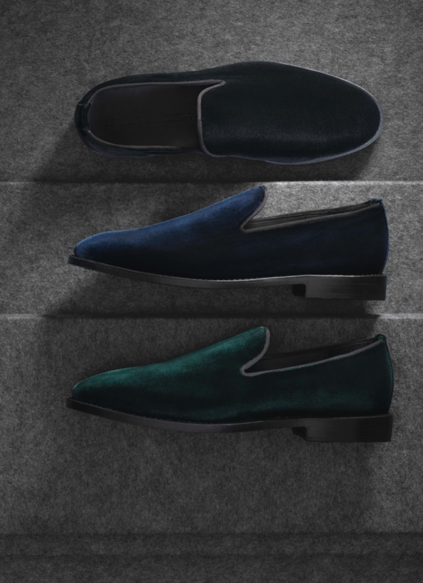A top view of three velvet loafers. One is black, one is blue, and one is green. The black one is displayed on its sole, while the green and blue shoes are displayed on their side.
