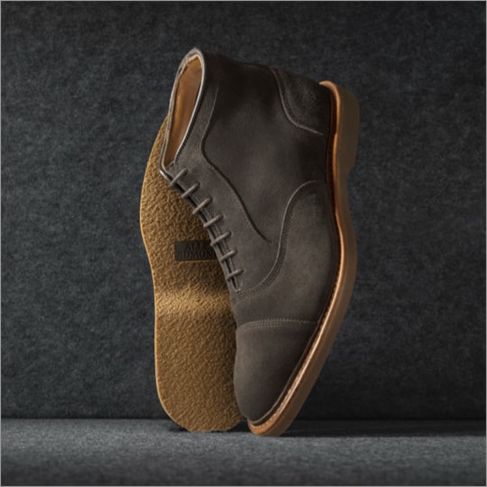 A pair of brown suede cap-toe oxford boots displayed vertically, with one sole facing forward.